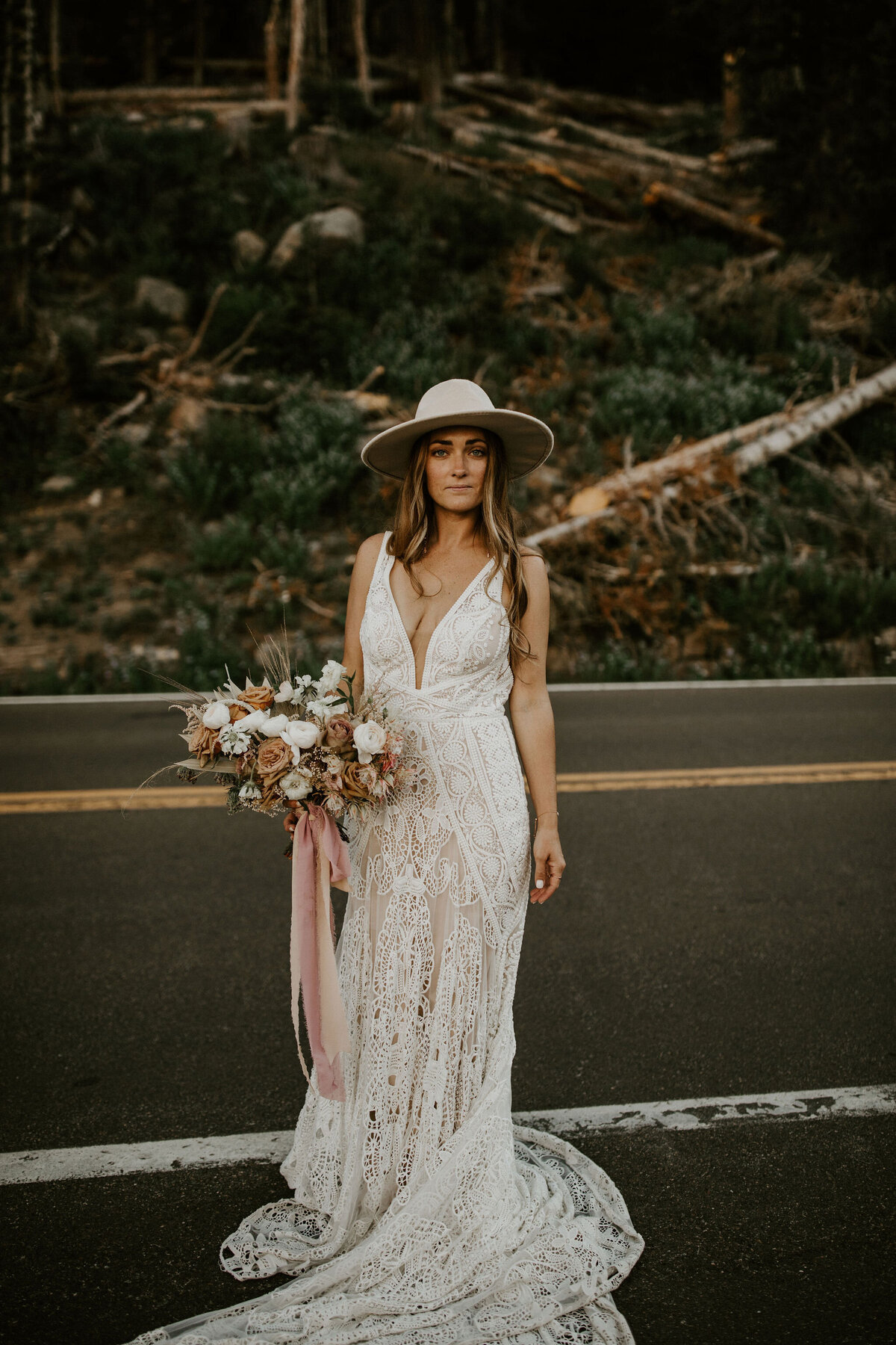 Bride wearing a white wedding gown and hat holding a bouquet of blush and white roses standing in the middle of the road.