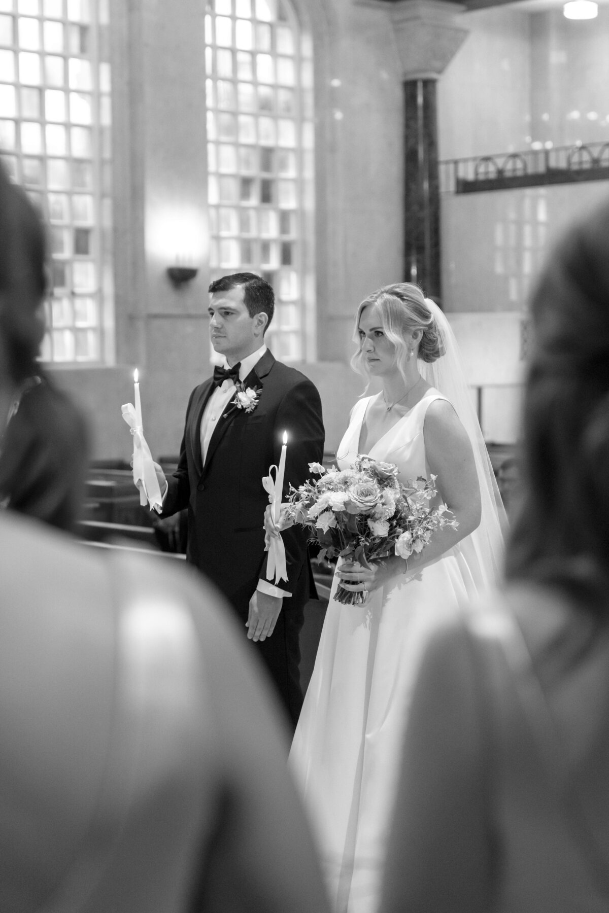 A bride and groom stand holding candles in a church. The bride wears a white dress and veil, holding a bouquet. The groom is in a suit with a bow tie. Theyre in an elegantly lit space, surrounded by blurred figures.