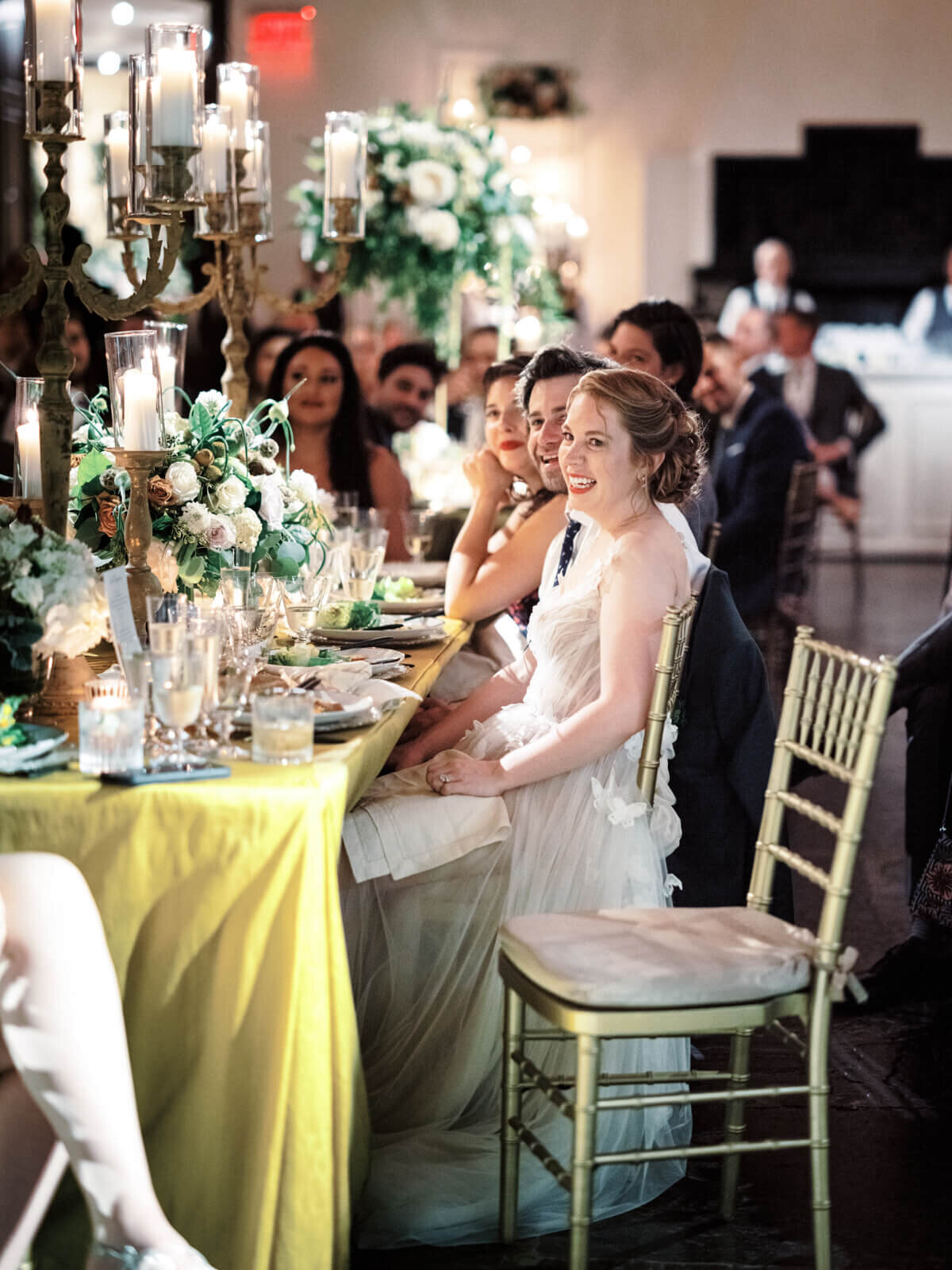 The bride is smiling with the guests while seated at a long dining table filled with flower and candle centerpieces.