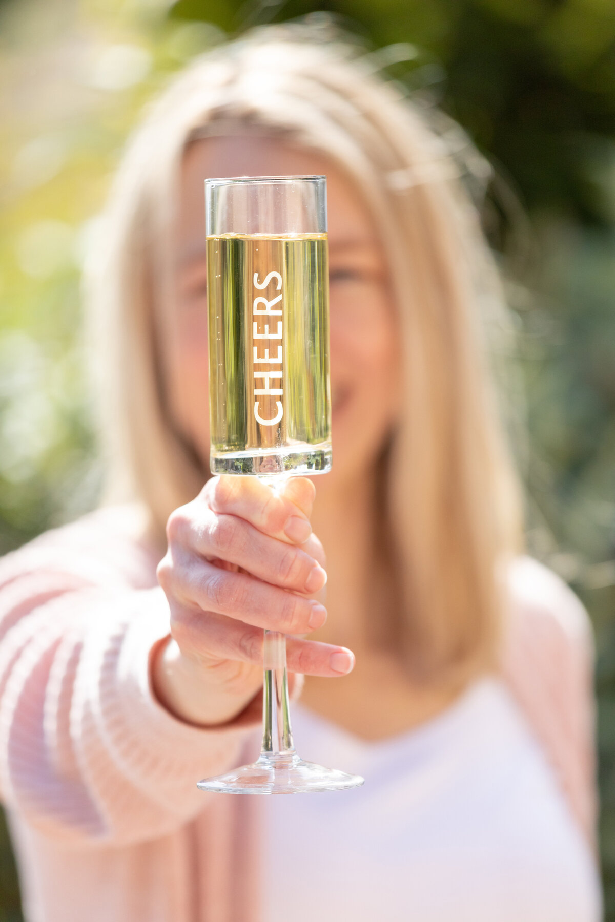 A smiling woman holds a champagne flute engraved with “Cheers” toward the camera, symbolizing celebration and confidence in a personal brand shoot for modern entrepreneurs.
