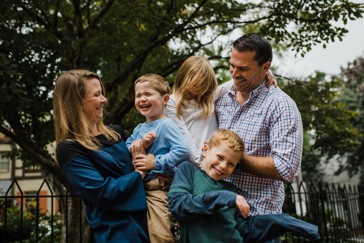 A couple and their three children sharing a smile outdoors in front of a large tree.