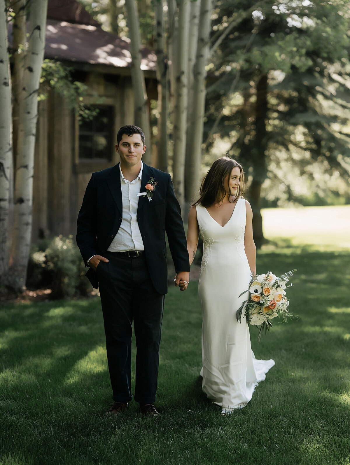 A beautiful couple, in an aspen colorado wedding, holds hands as the bride gazes off into the trees,
