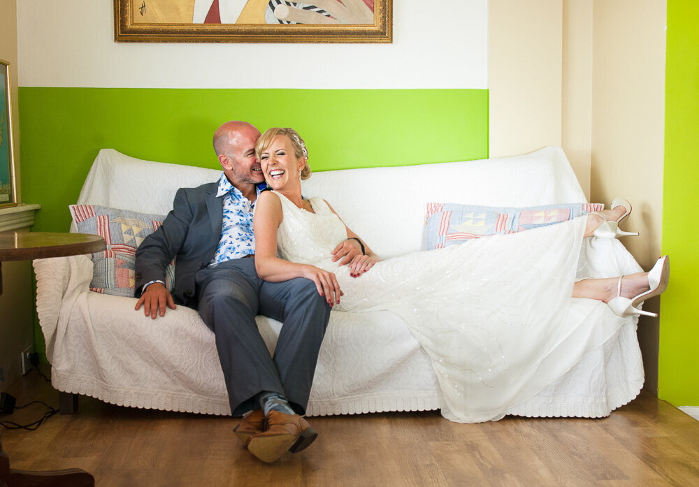 bride wearing a column-style wedding dress with straps sitting on a sofa with her groom wearing a blue suit and floral shirt in Greenes cafe, Inch beach