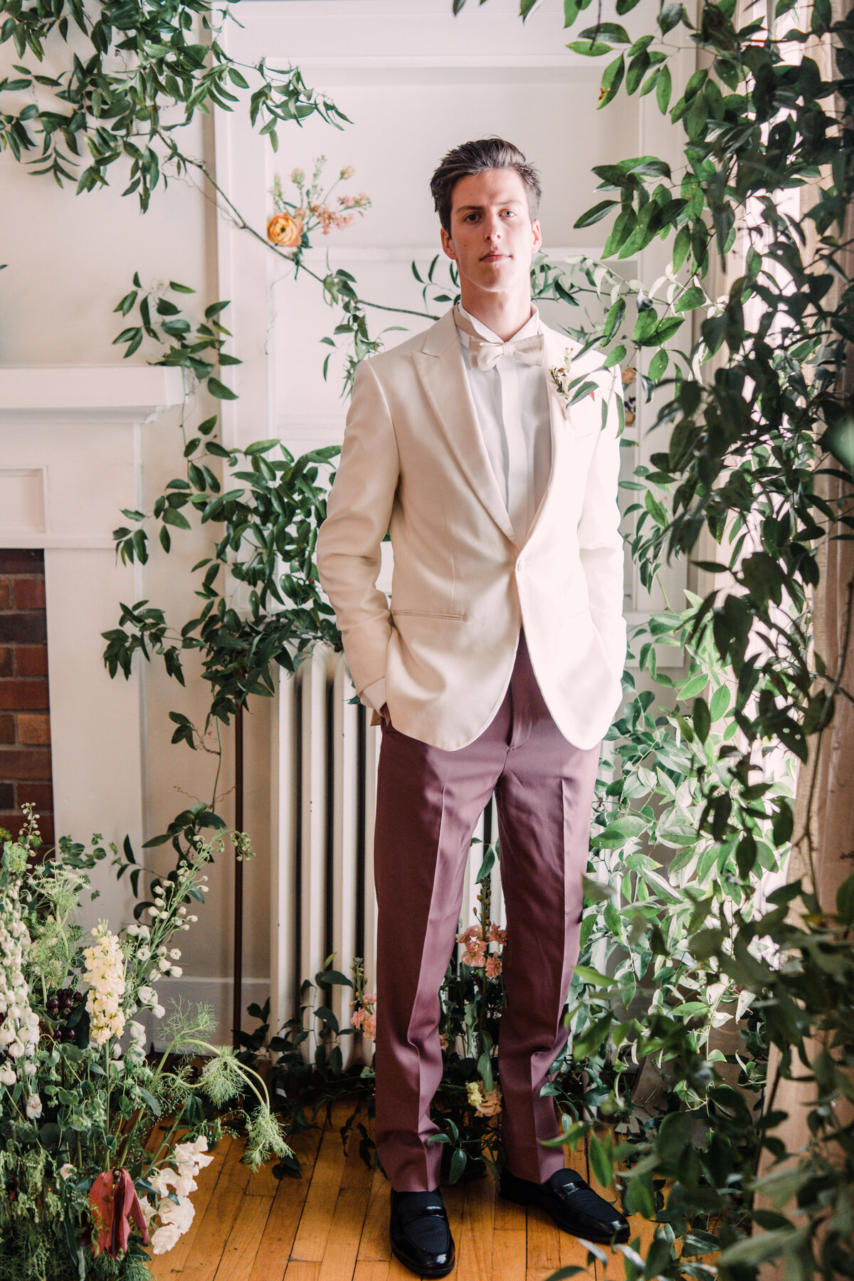 Groom wearing white tuxedo jacket & rose pants surrounded by vines & flowers in indoor garden