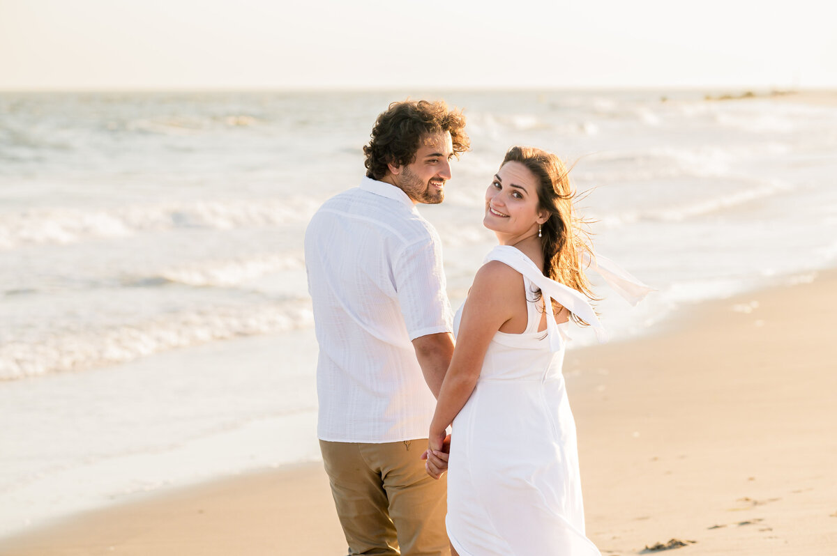 A man and woman hold hands and walk along a beach with waves in the background, both looking back and smiling.