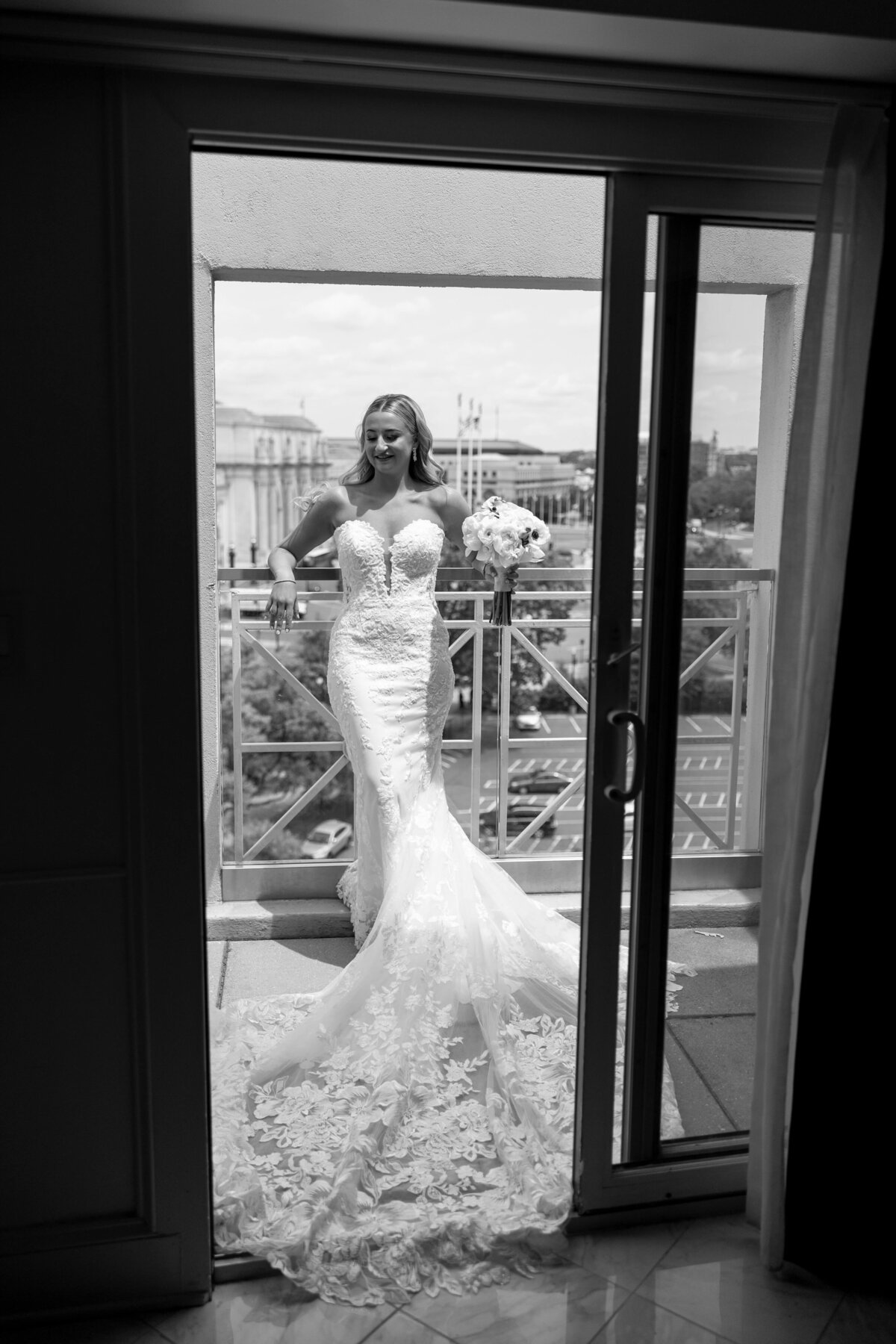 A bride standing on a balcony, wearing a long lace wedding dress. She holds a bouquet and looks out over a cityscape. The scene is captured in black and white.