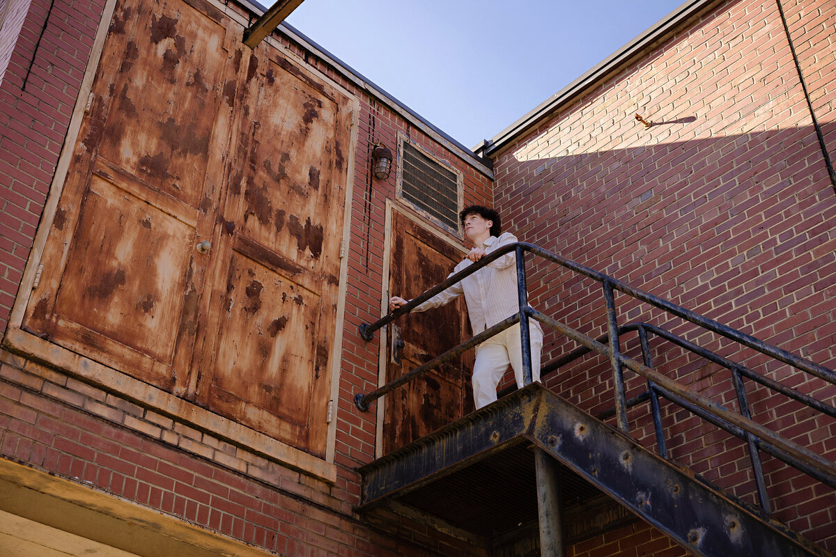 High school senior standing atop a tall staircase near a historic factory building at Camp North End, dressed in cream jeans and a striped shirt.