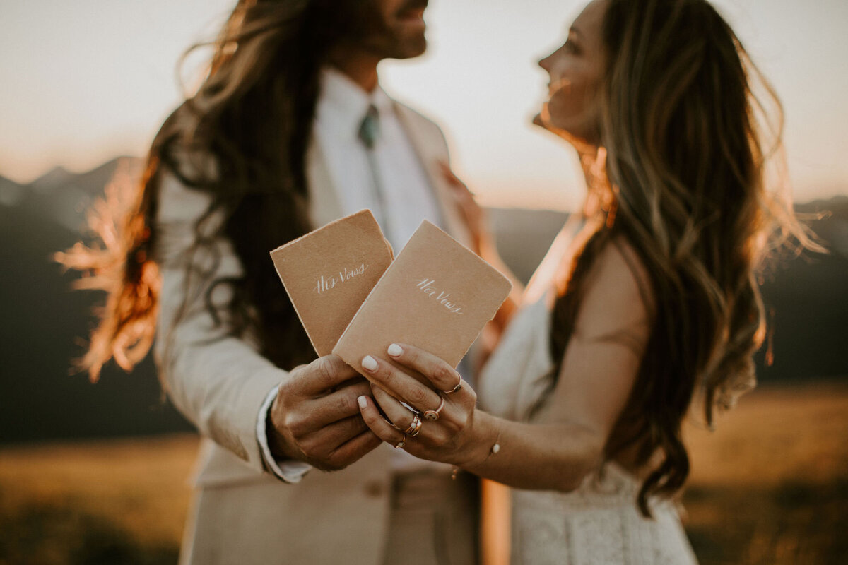 bride and groom pose facing each other holding tan and blush textured booklets with cursive white lettering