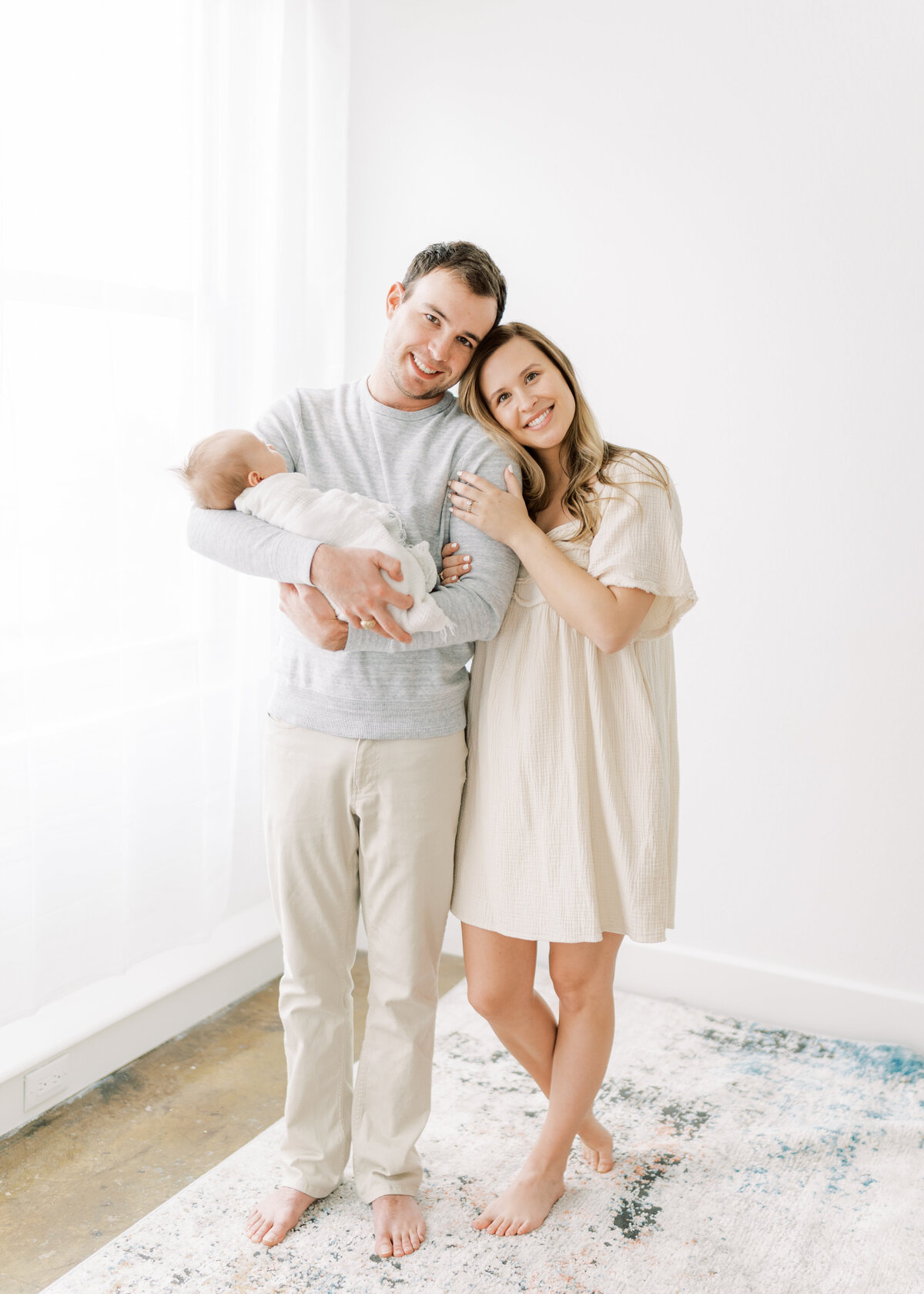 Portrait of a woman and a man wearing cream colored pants and a dress while holding a swaddled newborn baby atop a carpet