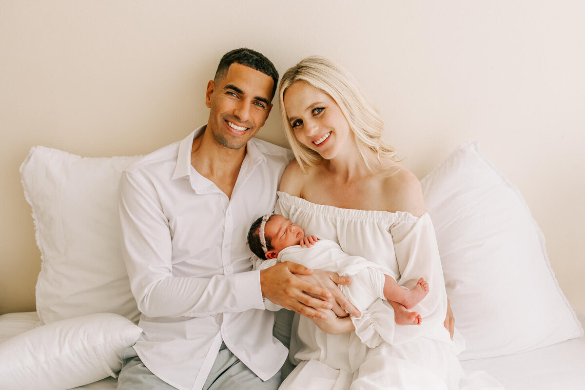 sweet happy family holding their newborn daughter and smiling. they are sitting on a bed in photographer Ann Marshall's studio