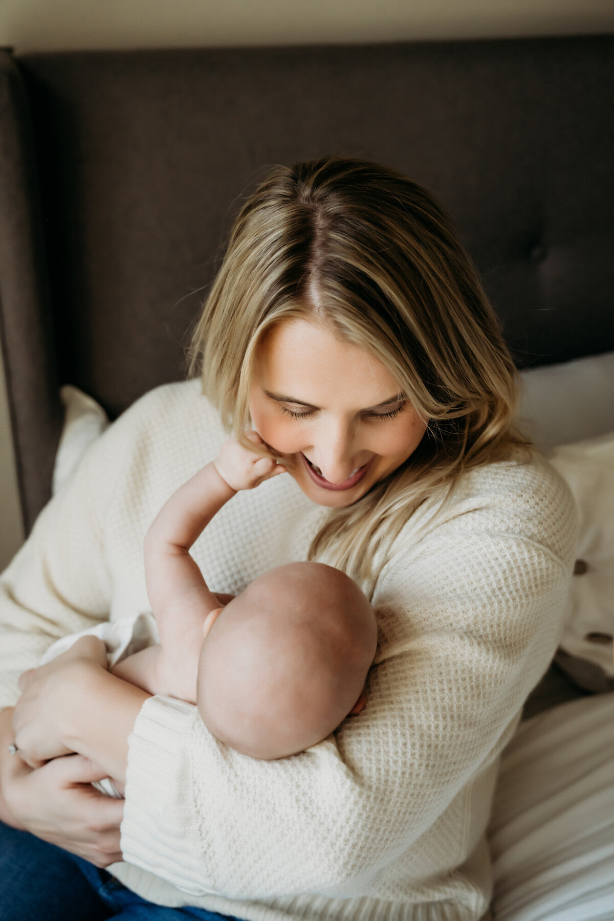 Newborn Photography, Mom is kissing baby and looking down at her, while wearing a white sweater and sitting on the bed. Baby's hand is on mom's face.