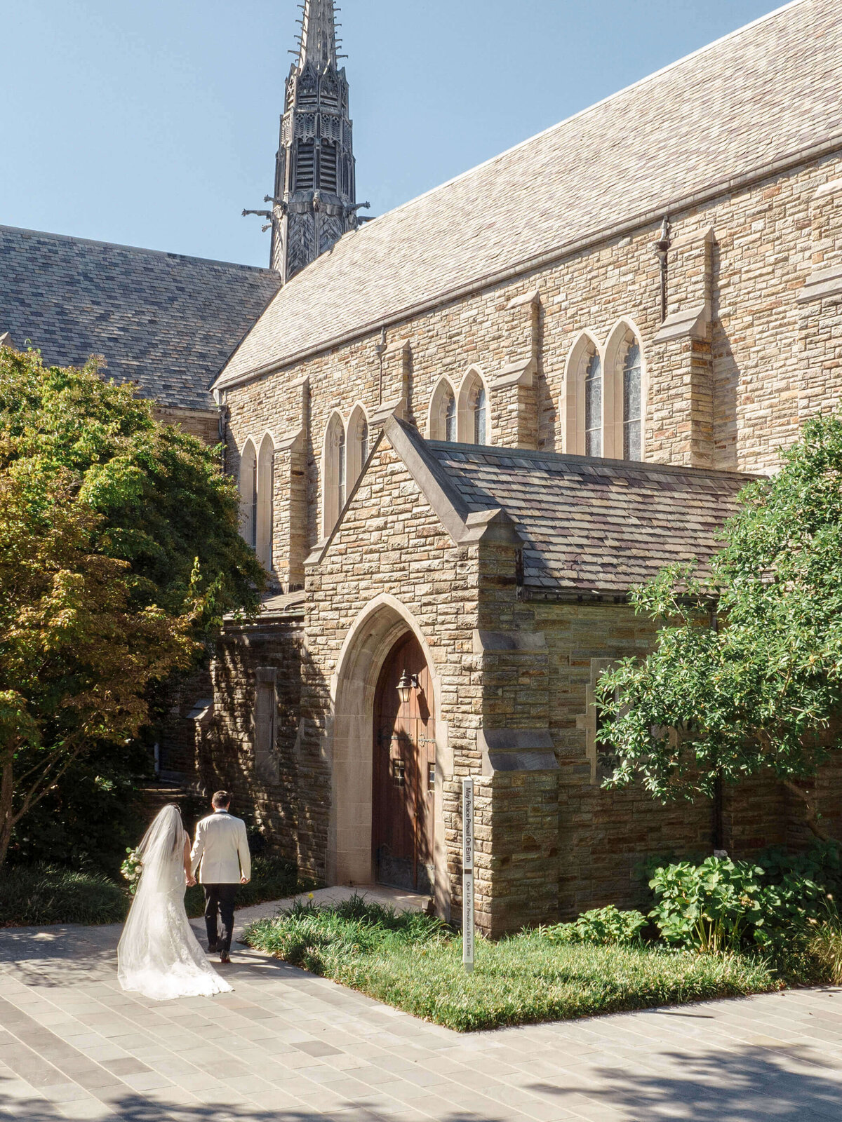 A bride and groom walk hand-in-hand toward a stone church with a tall steeple, surrounded by lush greenery on a sunny day. The bride wears a flowing white gown with a veil, and the groom is in a light-colored suit.
