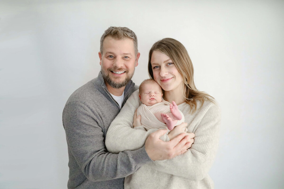 Parents smiling while holding wrapped newborn baby in light studio - Maple Grove photography