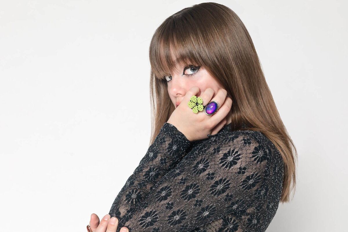 A woman with long brown hair stands against a white background. She is wearing a black lace dress and colorful rings on her fingers, with one hand raised to her mouth. Her expression is thoughtful, and she has bold eyeliner.