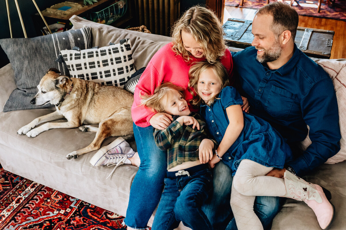 A family of four relaxes on a couch with their dog.