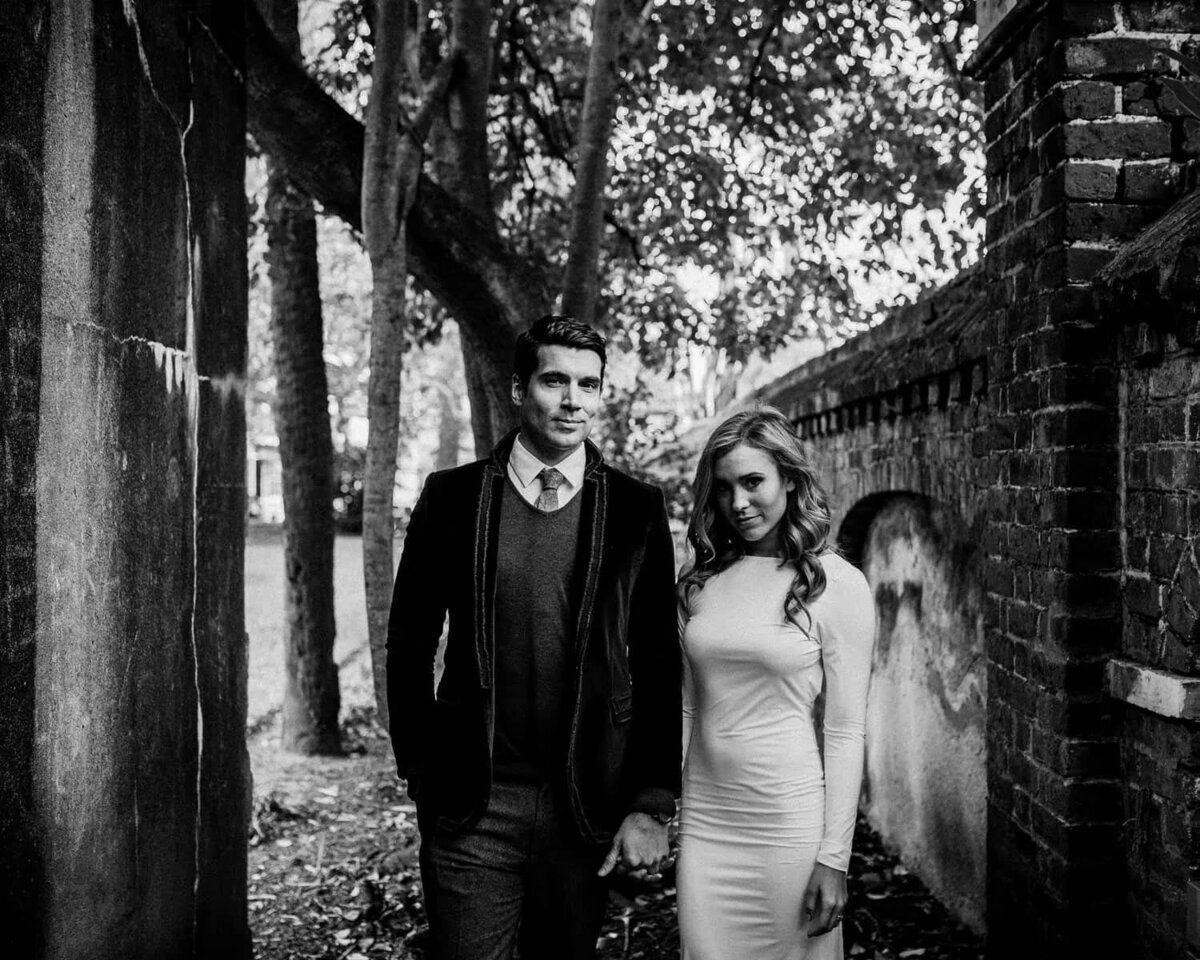 A wedding couple holding hands and standing next to a brink wall.
