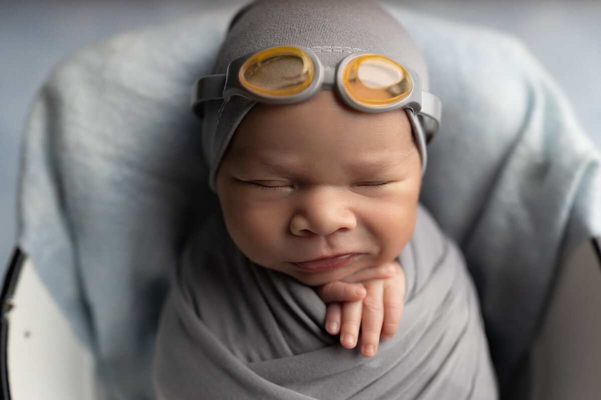 Fort Worth newborn photo of a swaddled baby wearing yellow swim goggles in a soft gray wrap, resting peacefully with a gentle expression.