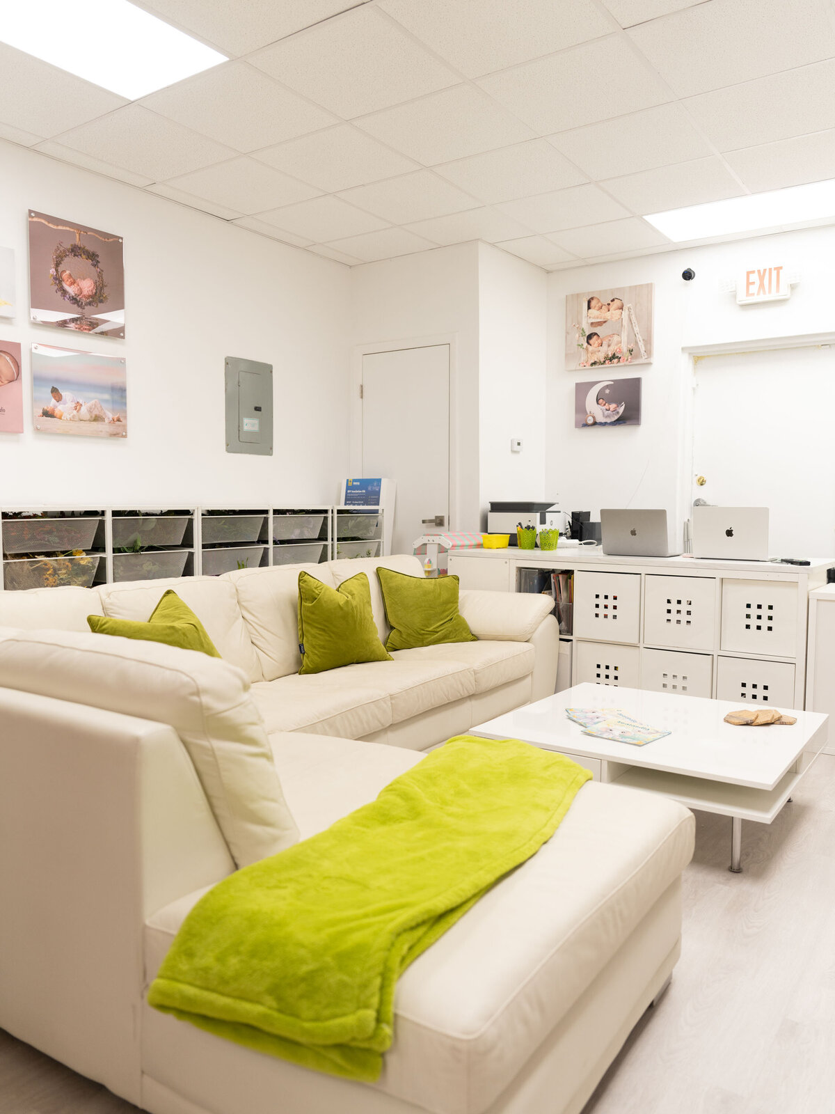 A cozy, well-lit room features a cream sectional sofa with green pillows and a matching throw. Artworks adorn the walls, and there's a modern white table.