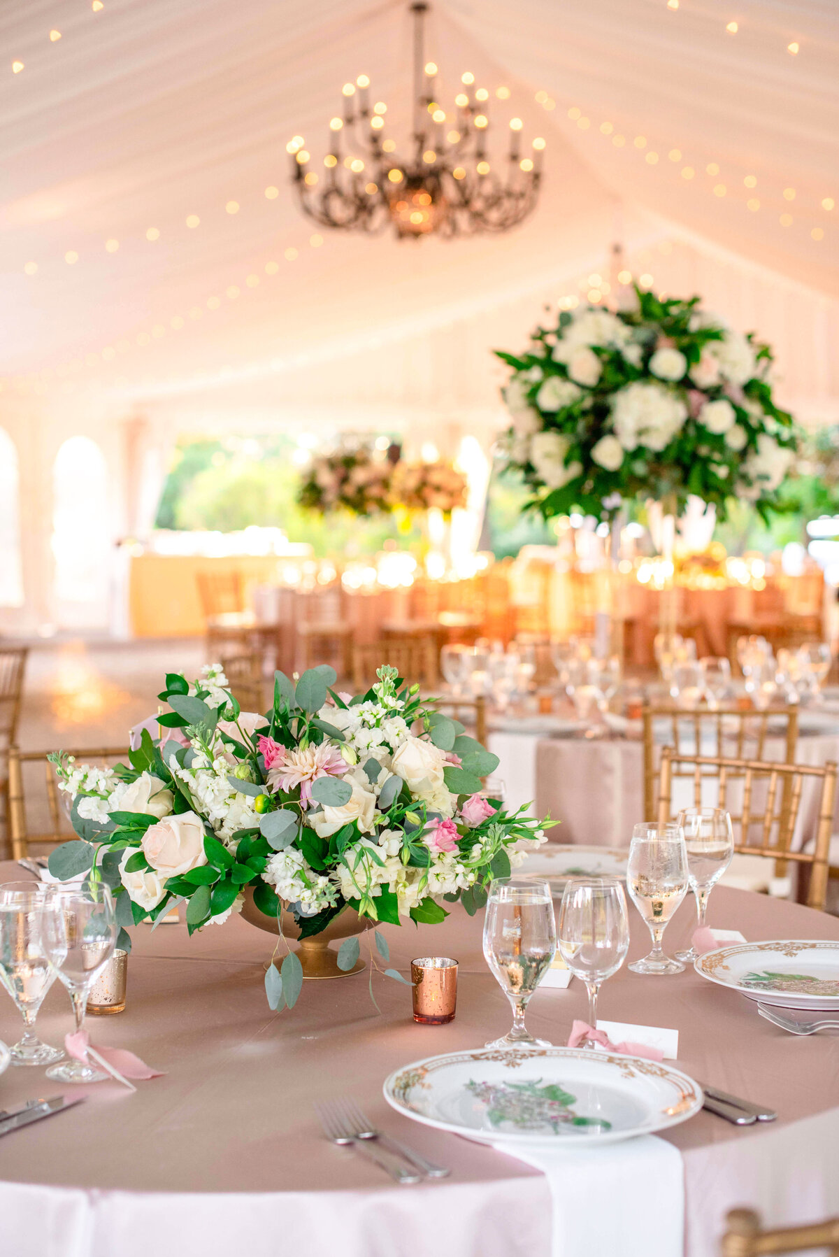 A beautifully decorated wedding reception room with elegant table settings. The tables feature floral centerpieces with white and pink flowers. A chandelier hangs from the ceiling, and soft lighting creates a warm ambiance.