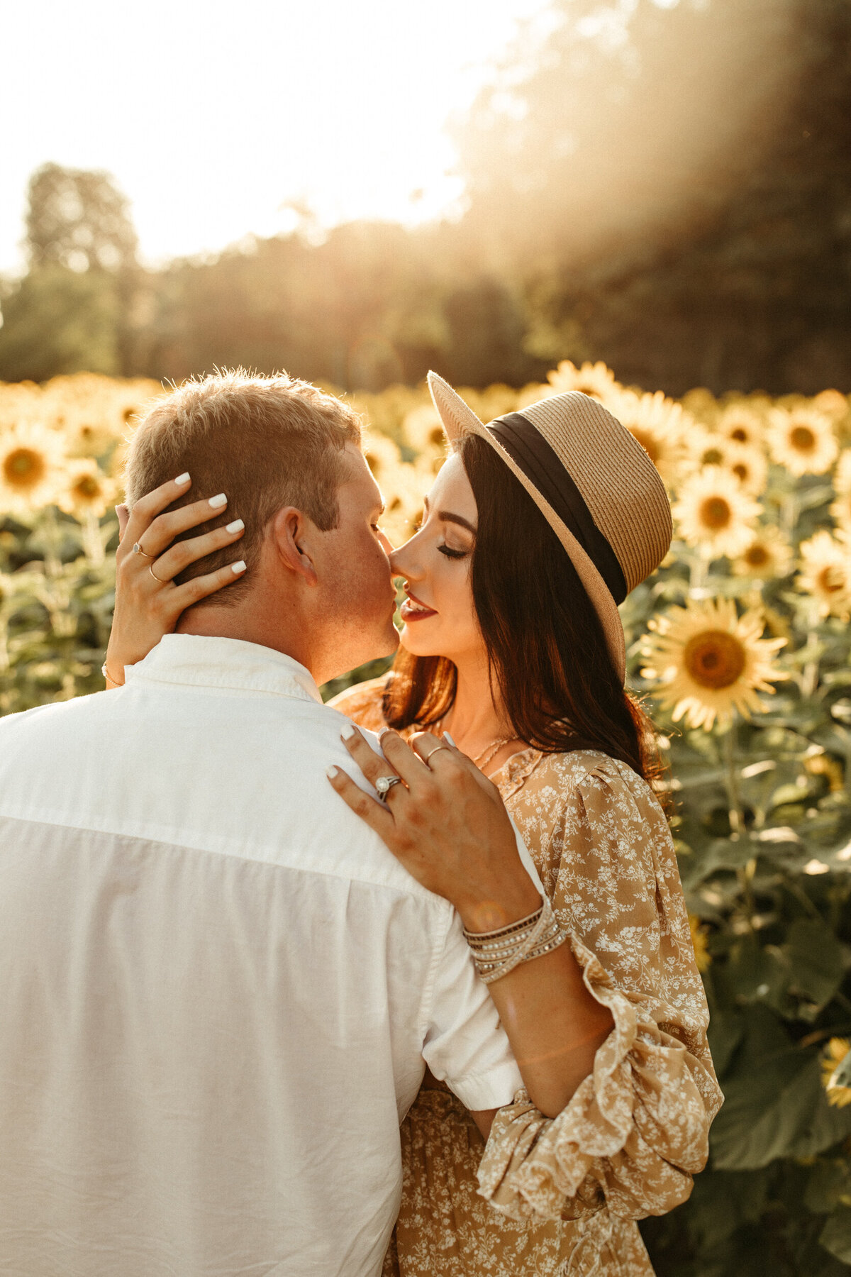 A girl in a yellow boho dress and hat is standing in a sunflower field at sunset and pulling in her fiancé for a kiss.