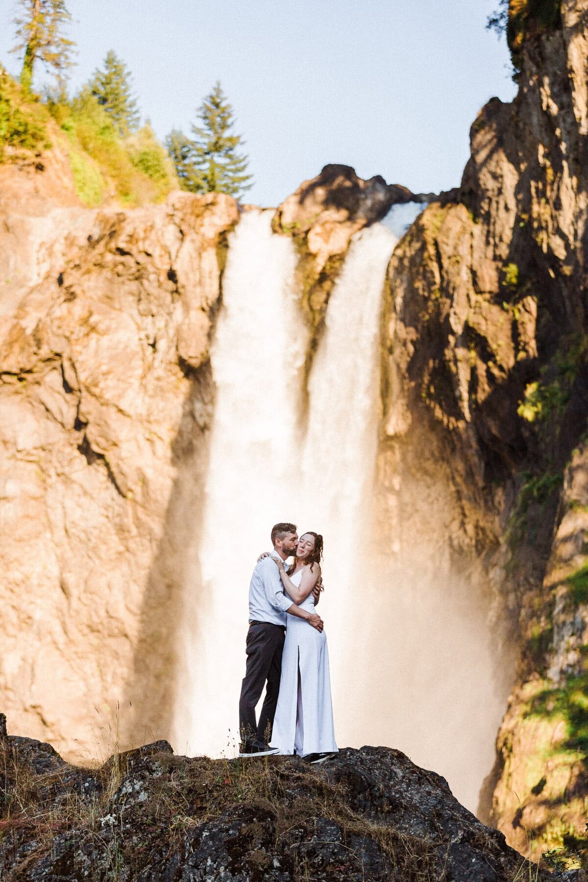 couple giving check kisses on top of rock in front of Snoqualmie Falls.