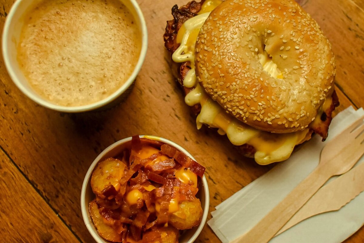 Bagel with meat and cheese sauce, with pot of loaded tater tots, a cup of coffee and a wooden knife and fork on a napkin. All shot from above