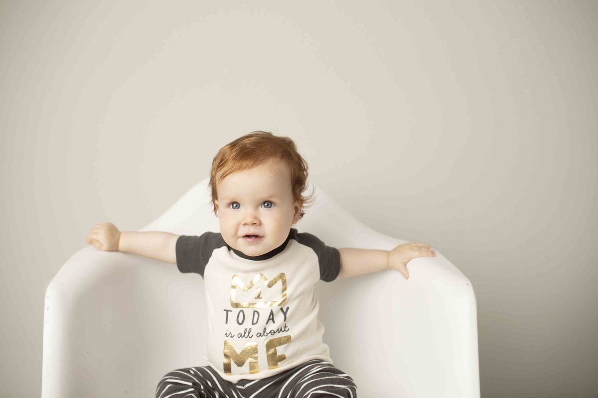 Smiling redhead birthday boy sitting comfortably in a white chair, enjoying his birthday photo session in the studio