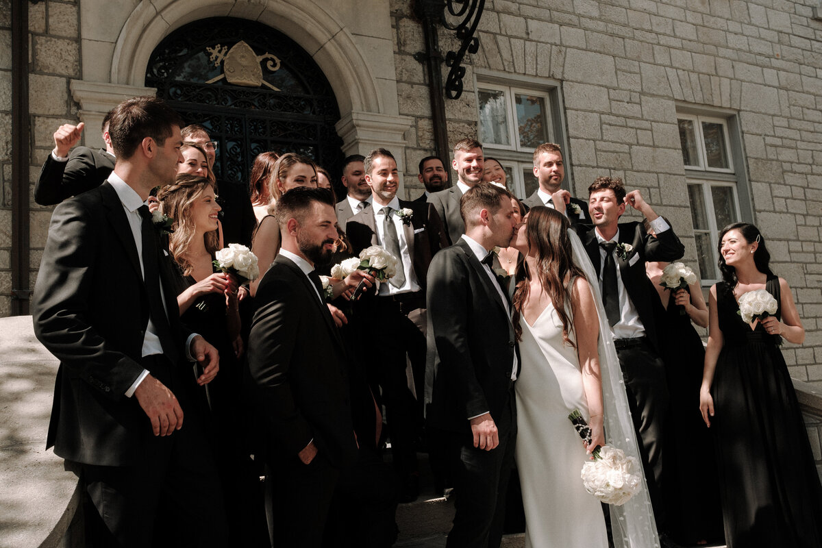 wedding party celebrating beside bride and groom kissing outside church