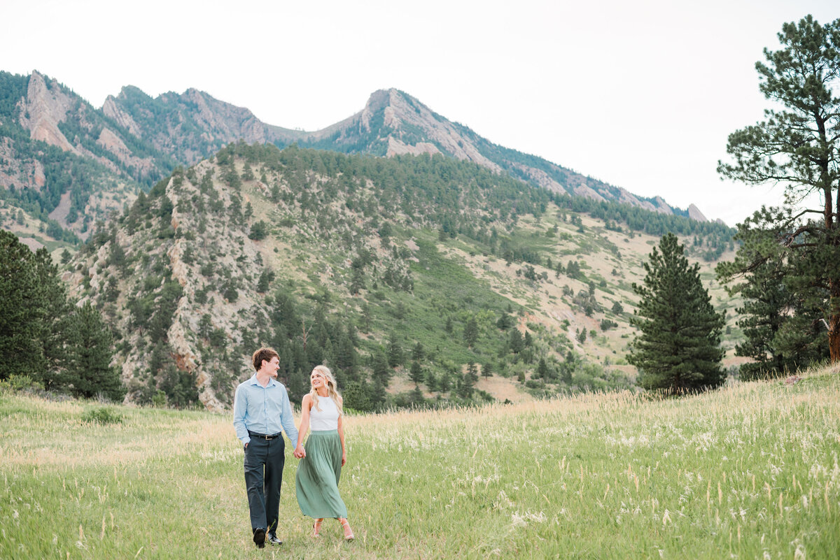 denver couples photography with denver wedding photographer capturing man and woman in formal attire walking through a field while holding hands and looking to one another with the Rocky Mountains in the distance