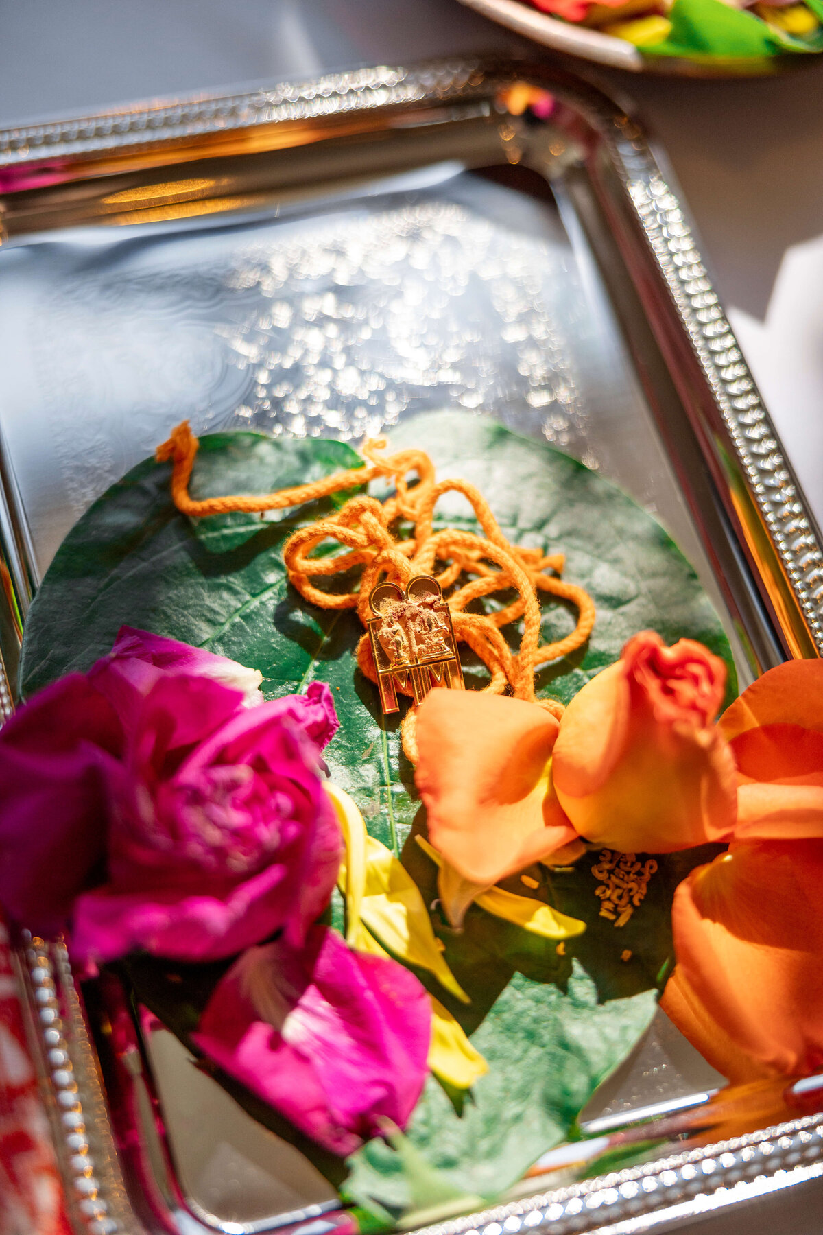 A gold necklace with a pendant rests on a green leaf, surrounded by vibrant orange and pink flowers, placed on a decorative silver tray.