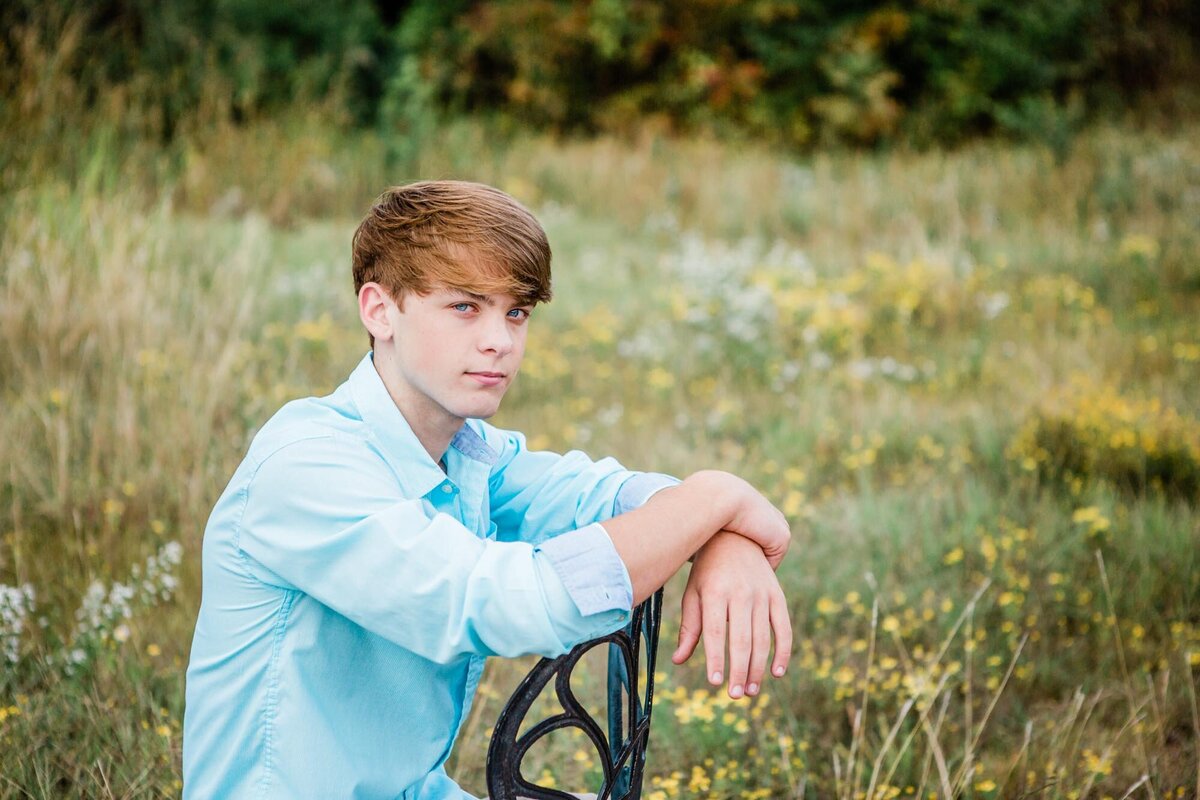 Senior portrait of a boy with striking blue eyes sitting backward in a chair in a  field