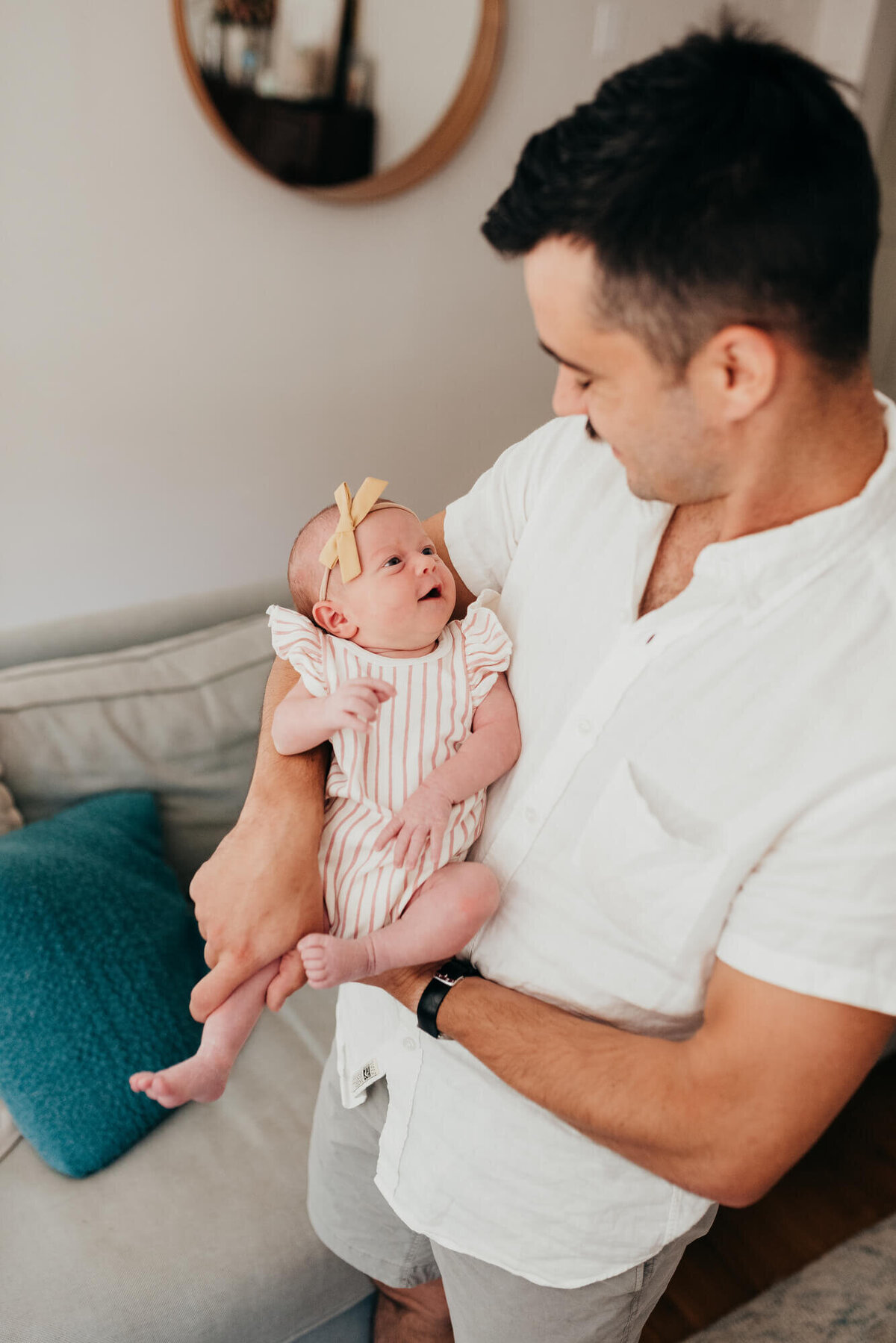 A young father wearing a bright white shirt holds his newborn daughter, she is wearing a cute white and pink striped romper