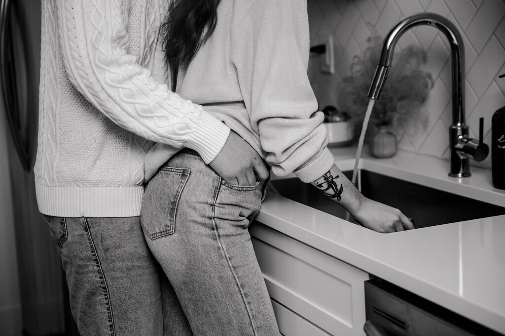 husband holding wife's hips while washing dishes in black and white