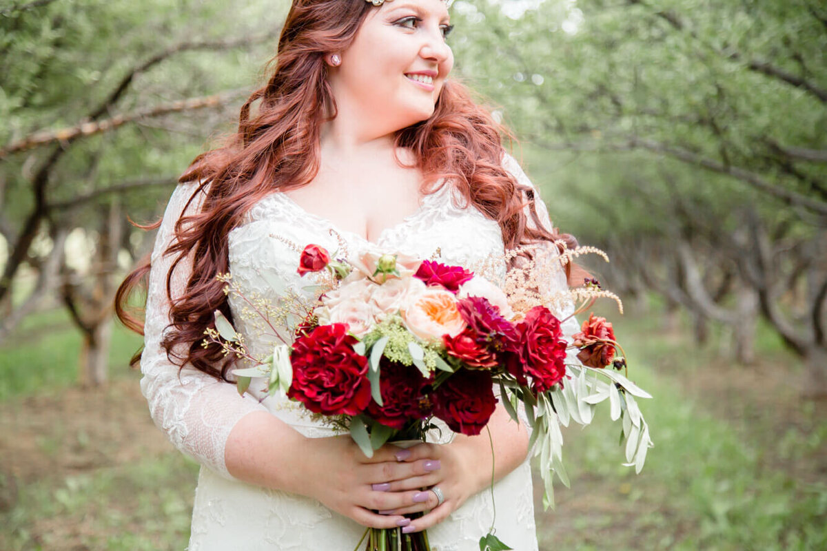 redheaded bride holding a bouquet of red and white roses in an apple orchard