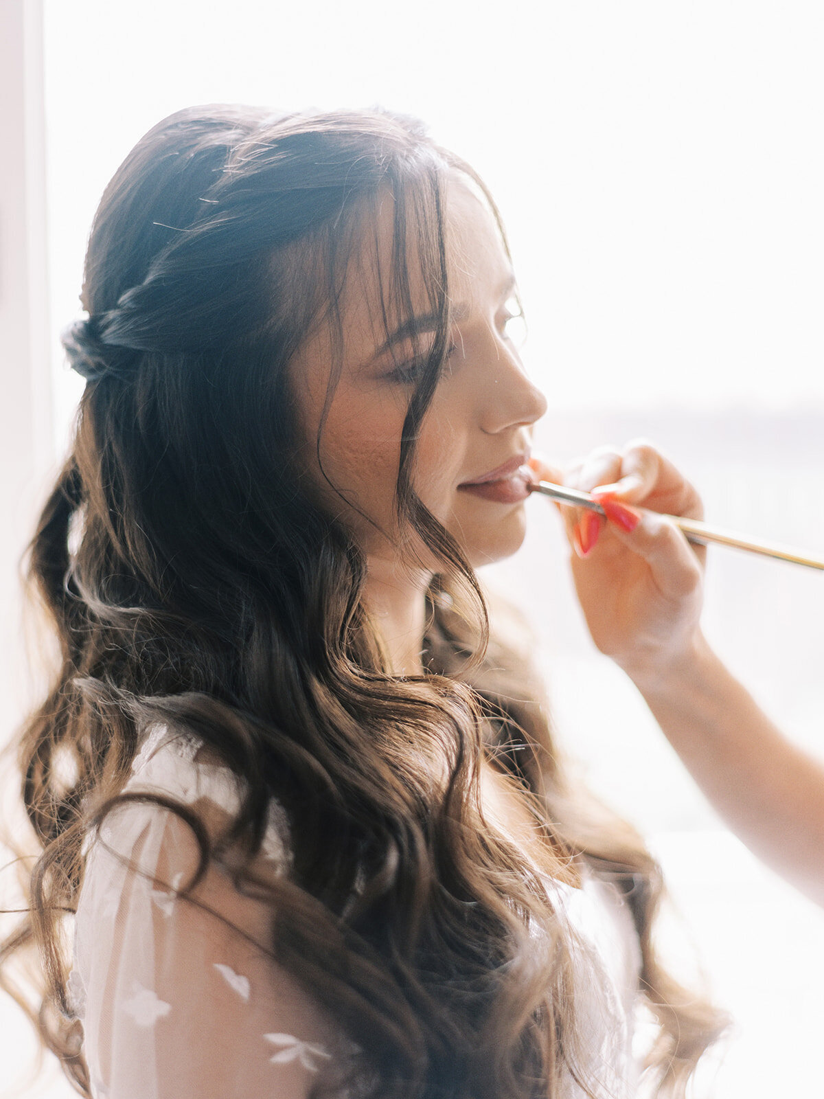 A woman with long, wavy hair is having lipstick applied by another person using a makeup brush. She is sitting near a light source, preparing for a classic Calgary wedding at Fairmont Palliser.