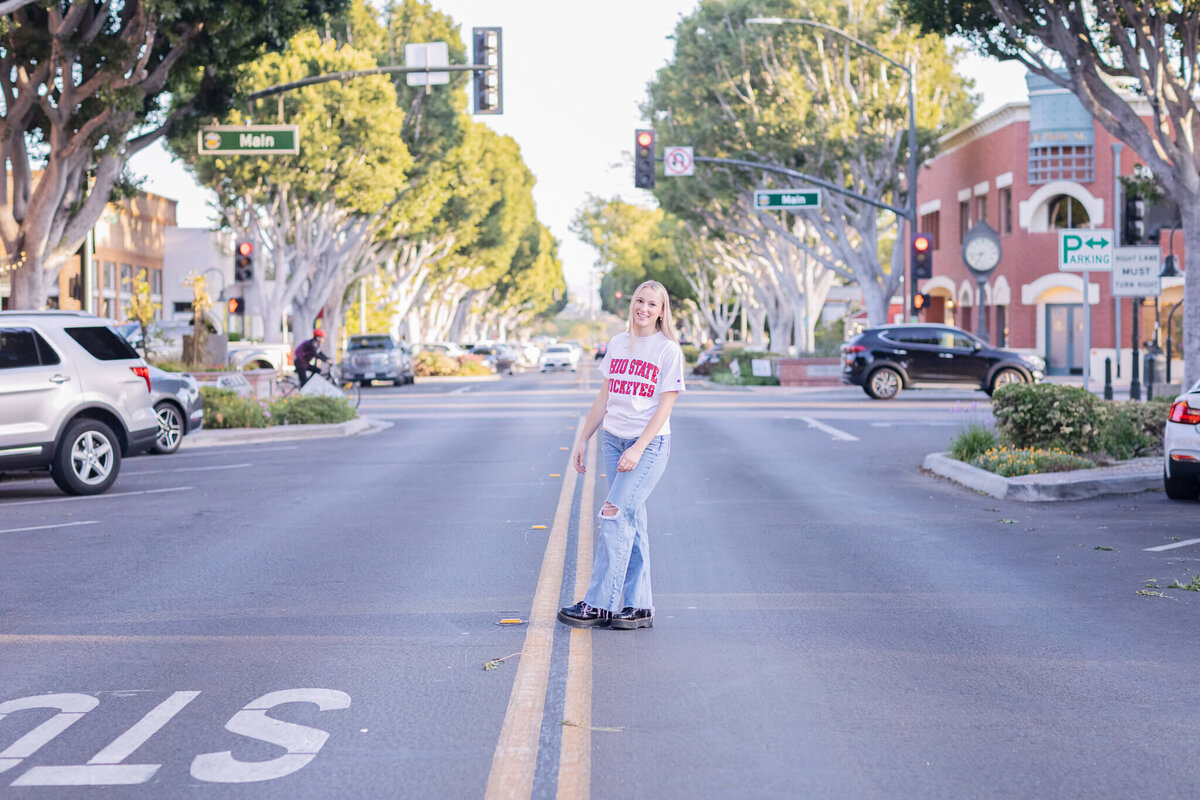 A person stands in the middle of a quiet street lined with parked cars and trees. They wear a white shirt and jeans, smiling and facing the camera. The background shows buildings and traffic lights.