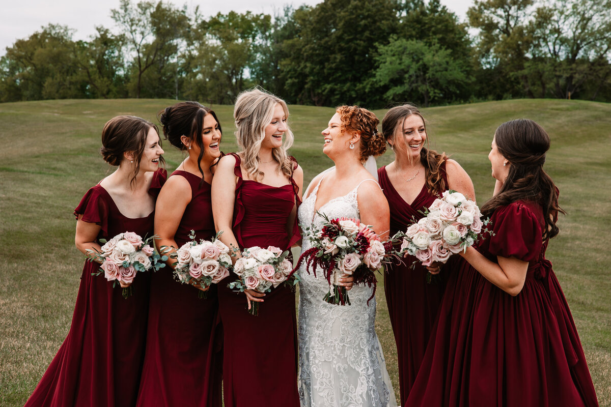 A bride laughs with her bridesmaids in red gowns in a field holding their matching bouquets