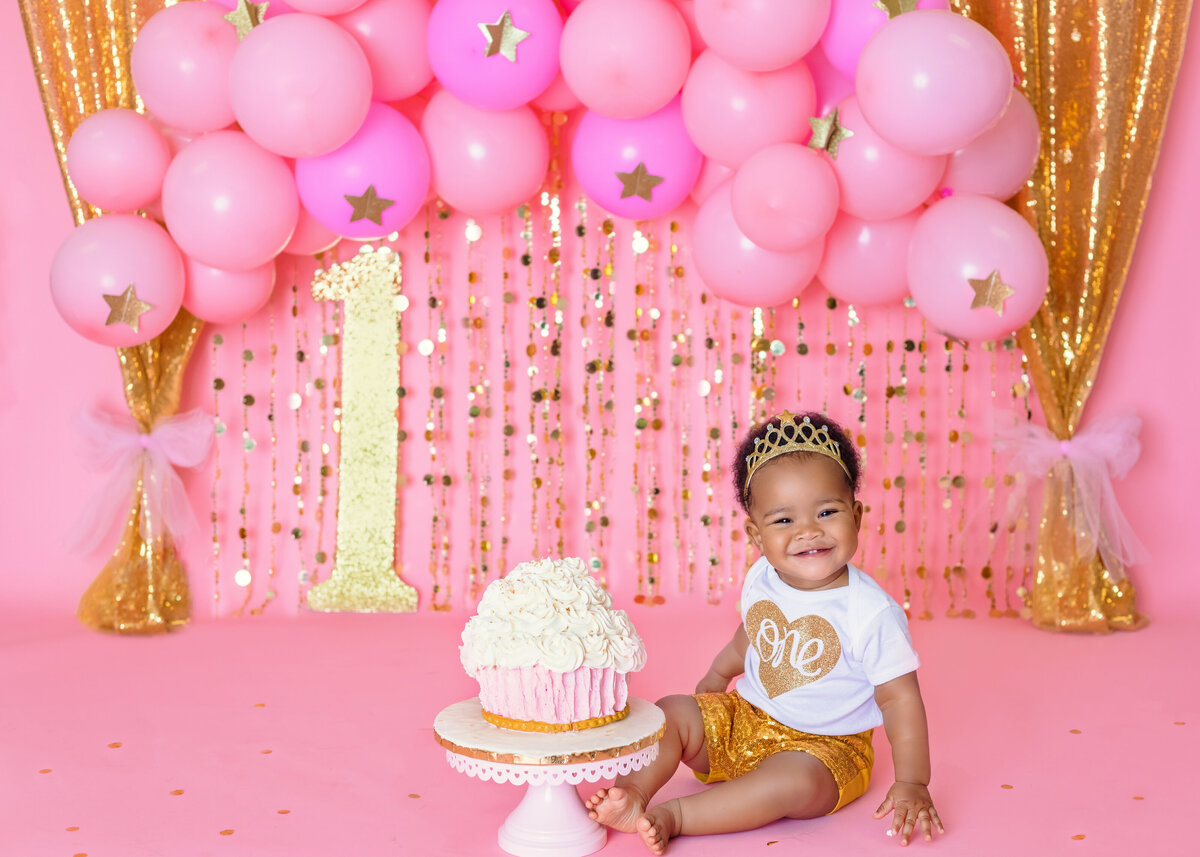 Cake Smash Photographer, a baby girl smiles before her large cupcake shaped cake before balloons