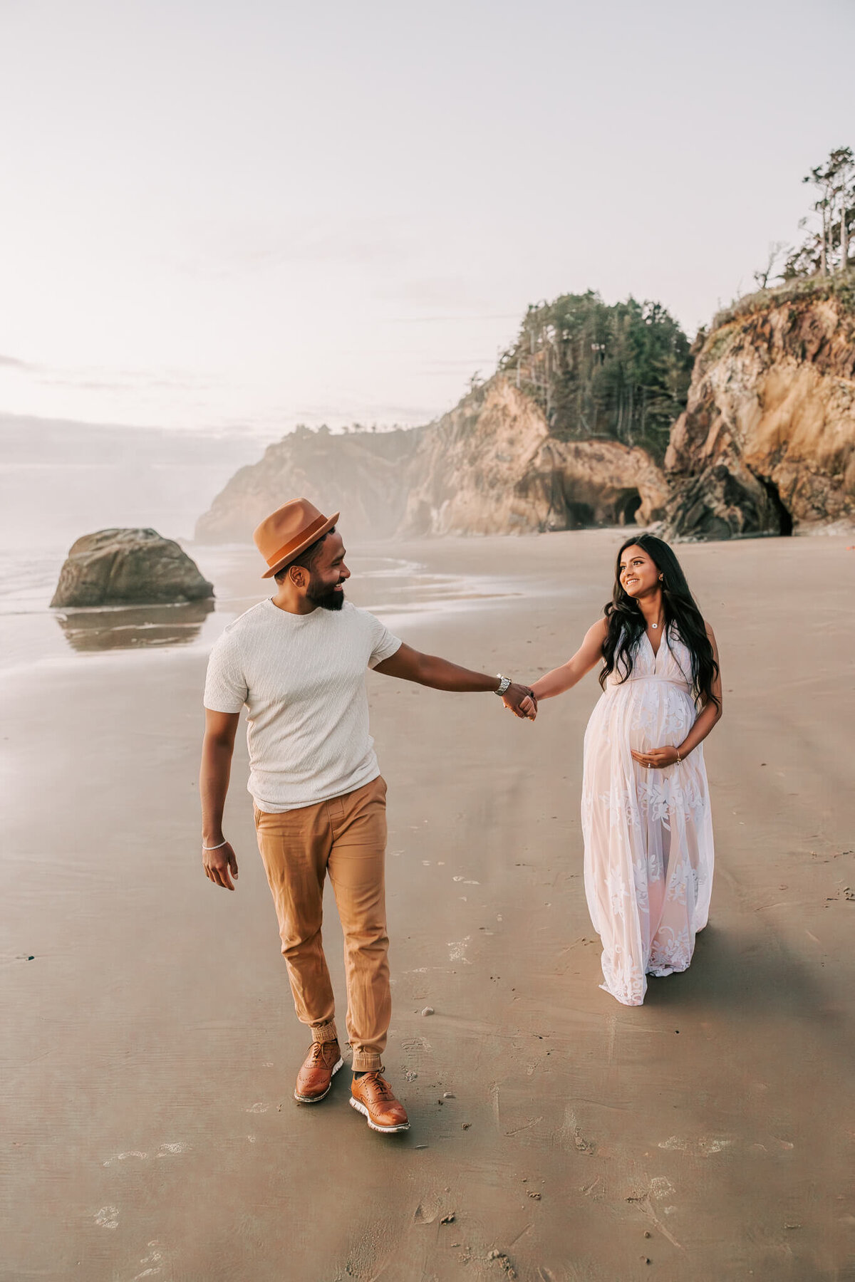 Portrait of a beautiful couple walking holding hands and smiling. She has her hand on her pregnant belly and is wearing a white dress. She has long black hair and he is wearing a tan hat and slacks.
