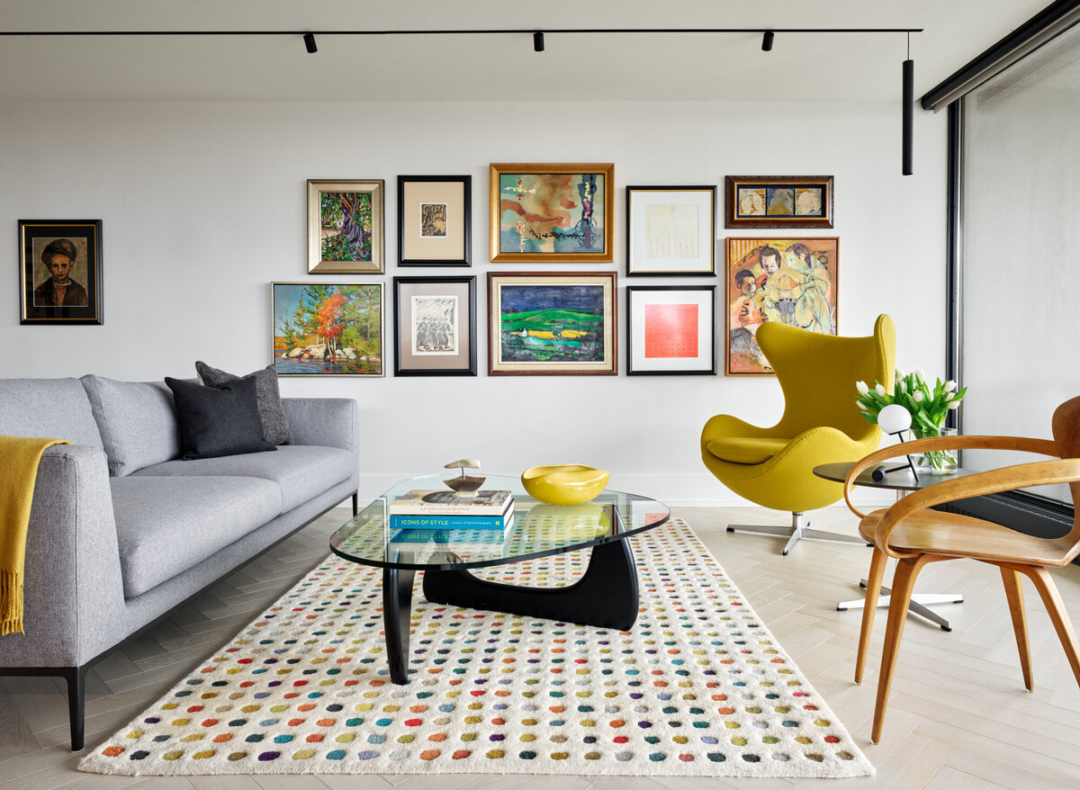 A bright, spacious living room featuring stylish grey, yellow, and brown seats, a glass coffee table, colourful patterned rug, and various framed art pieces on the wall