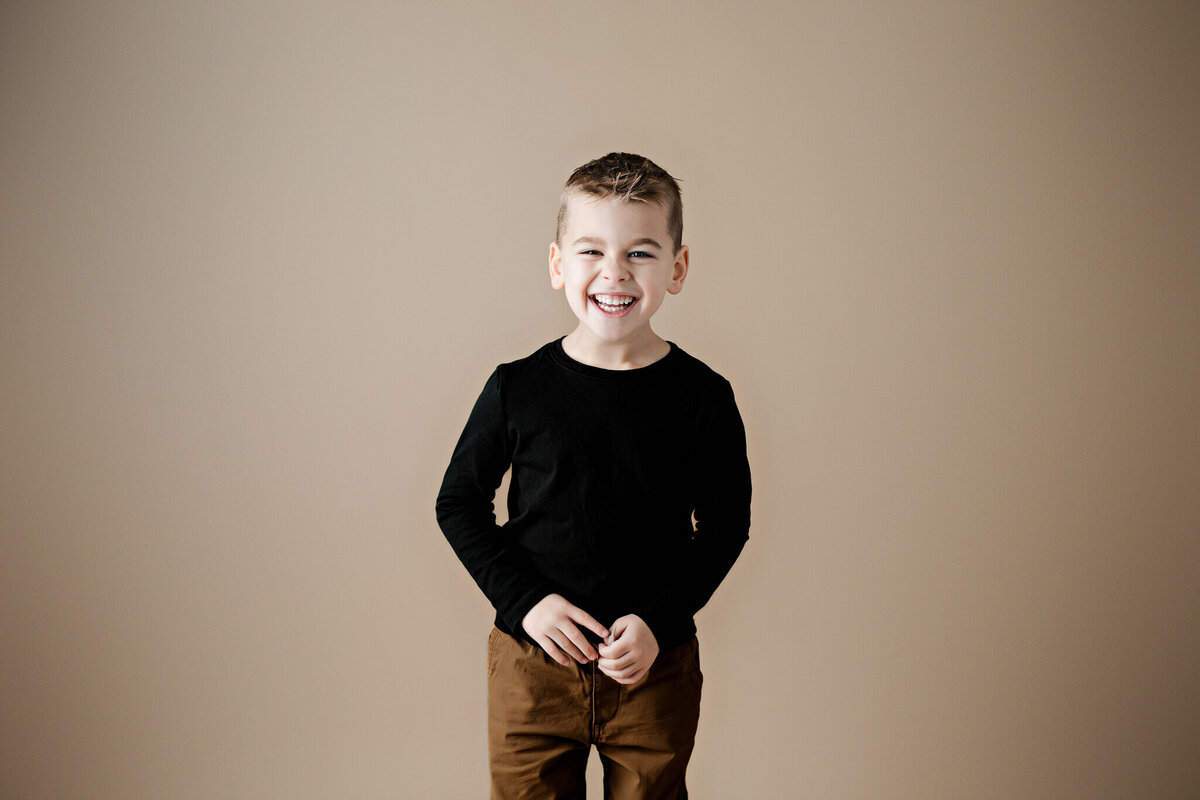 Smiling young boy with short hair, barefoot and wearing a black long-sleeve shirt and brown pants, stands against a beige background with his hands in his pockets—a perfect milestone celebration moment for any Bloomington cake smash photographer to capture.