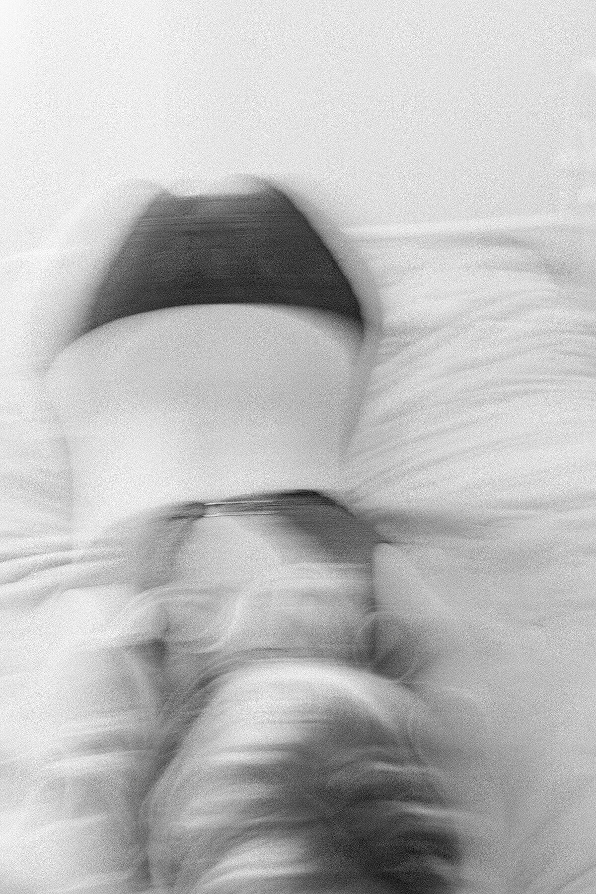 A blurred black and white photo of a woman leaning on a bed while wearing black lace lingerie.