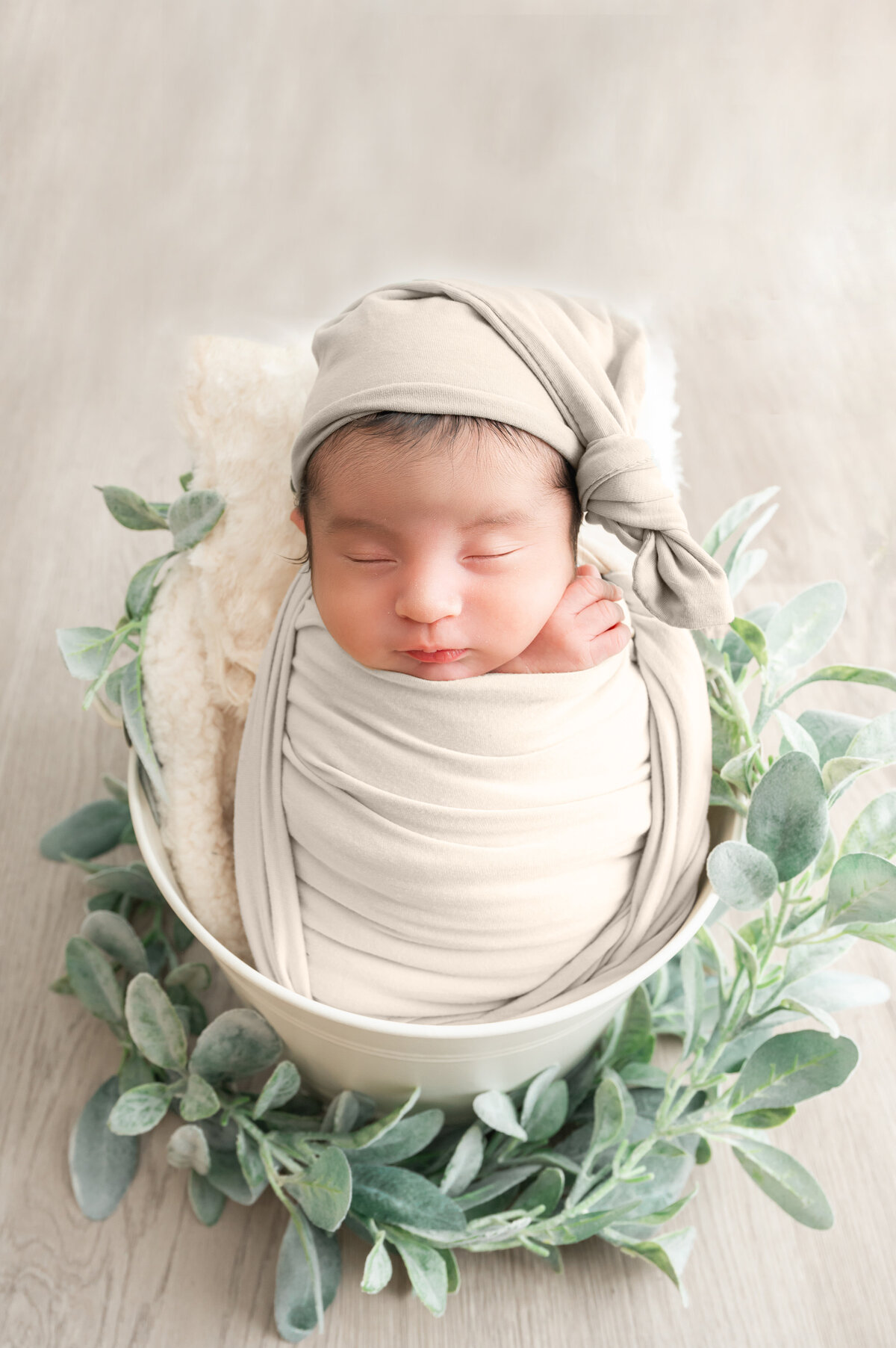 Newborn swaddled in off-white wrap and bonnet, nestled in a bucket surrounded with greenery.