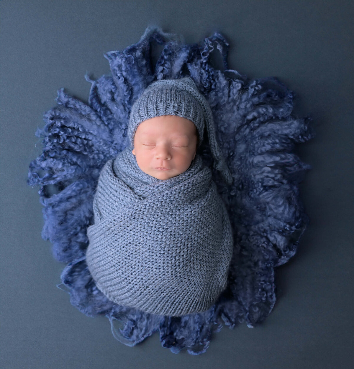 Newborn wrapped in blue knitted wraps and matching hat sleeping on a bed of denim colored blue curly blanket on solid blue backdrop. Captured by Beth Benecki of E.Benecki Photography.