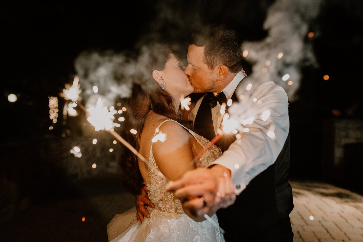 A bride and groom kissing while holding out sparklers.