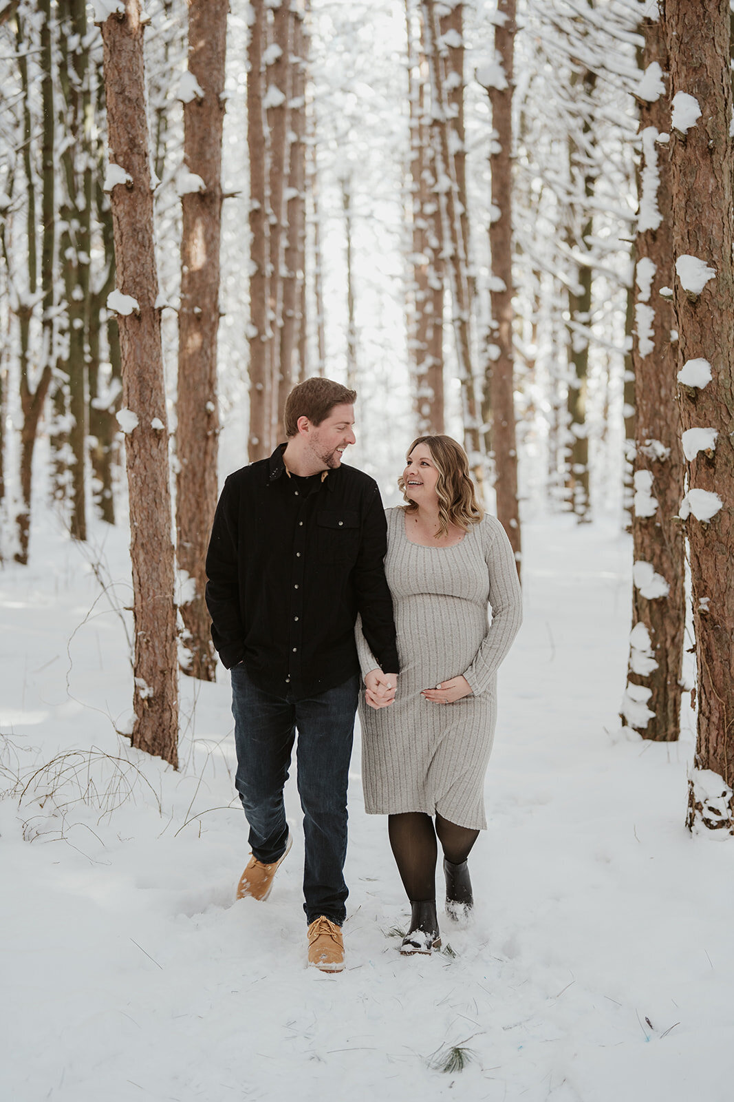 A pregnant mom and dad walking through the snowy pines at Al sabo Preserve in Kalamzoo, MI
