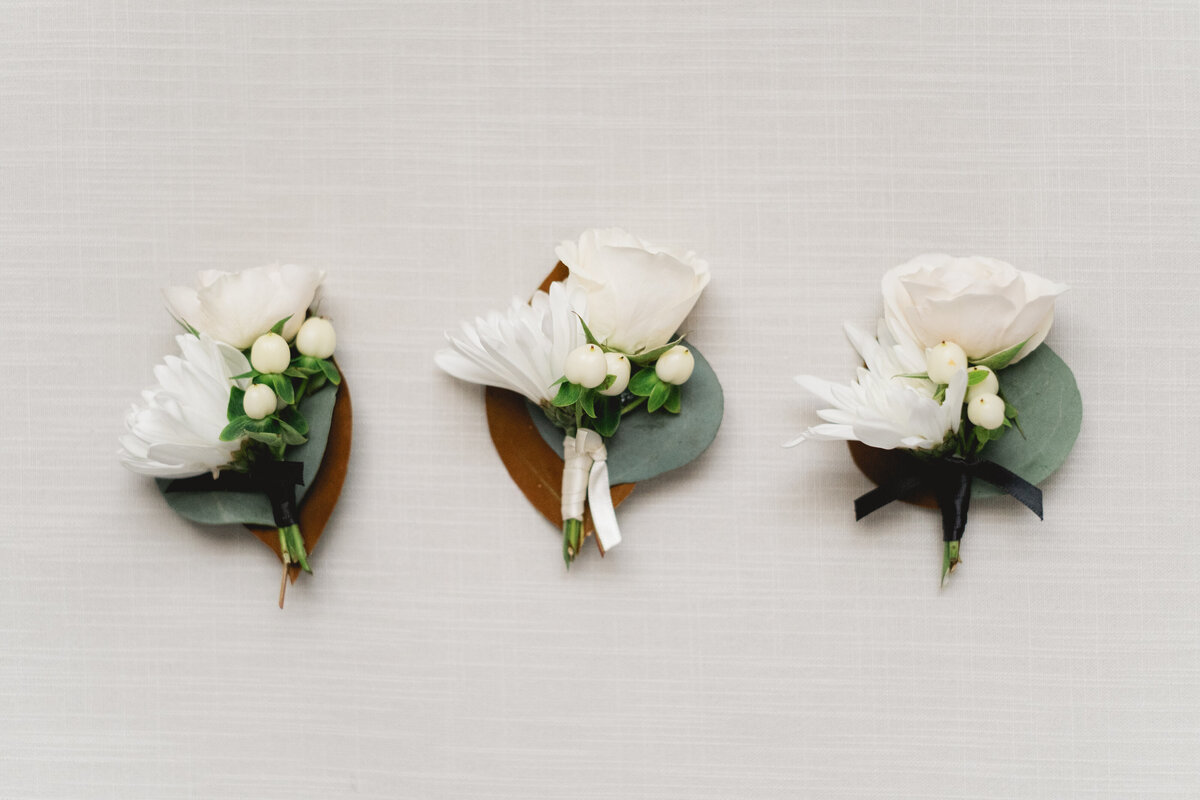 three white flower boutonnieres sitting on white linen backdrop