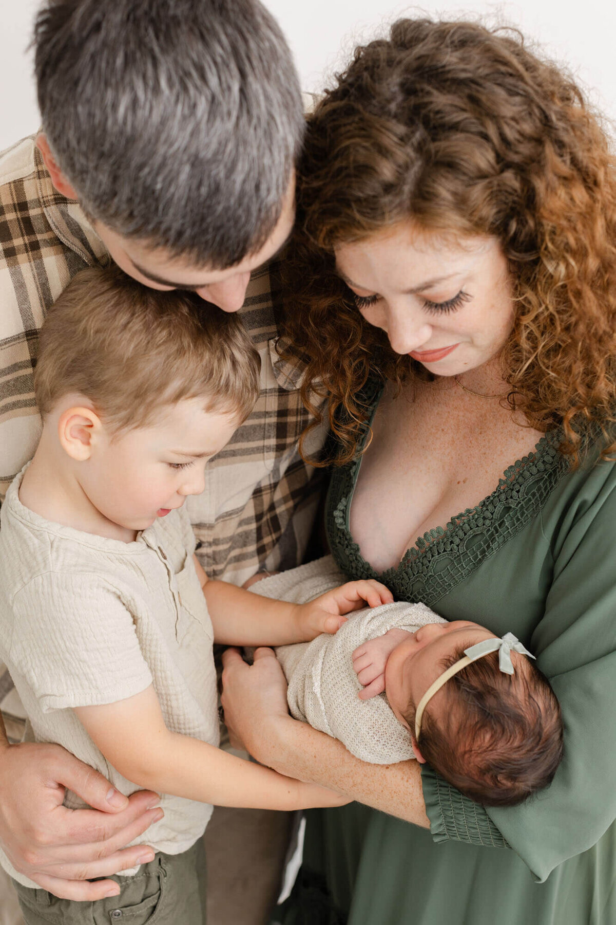 Mom and dad in greens and beiges are standing next to toddler son and mom is holding newborn baby. All family members are looking down at baby.