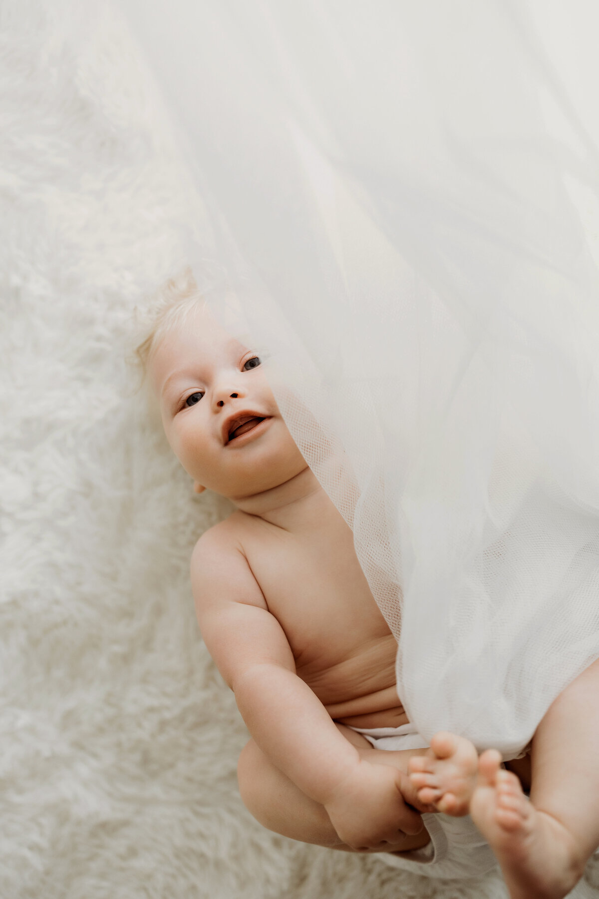 Happy baby boy wearing a only a white diaper cover lays on his back and plays with white sheer curtains during his Denver milestone session