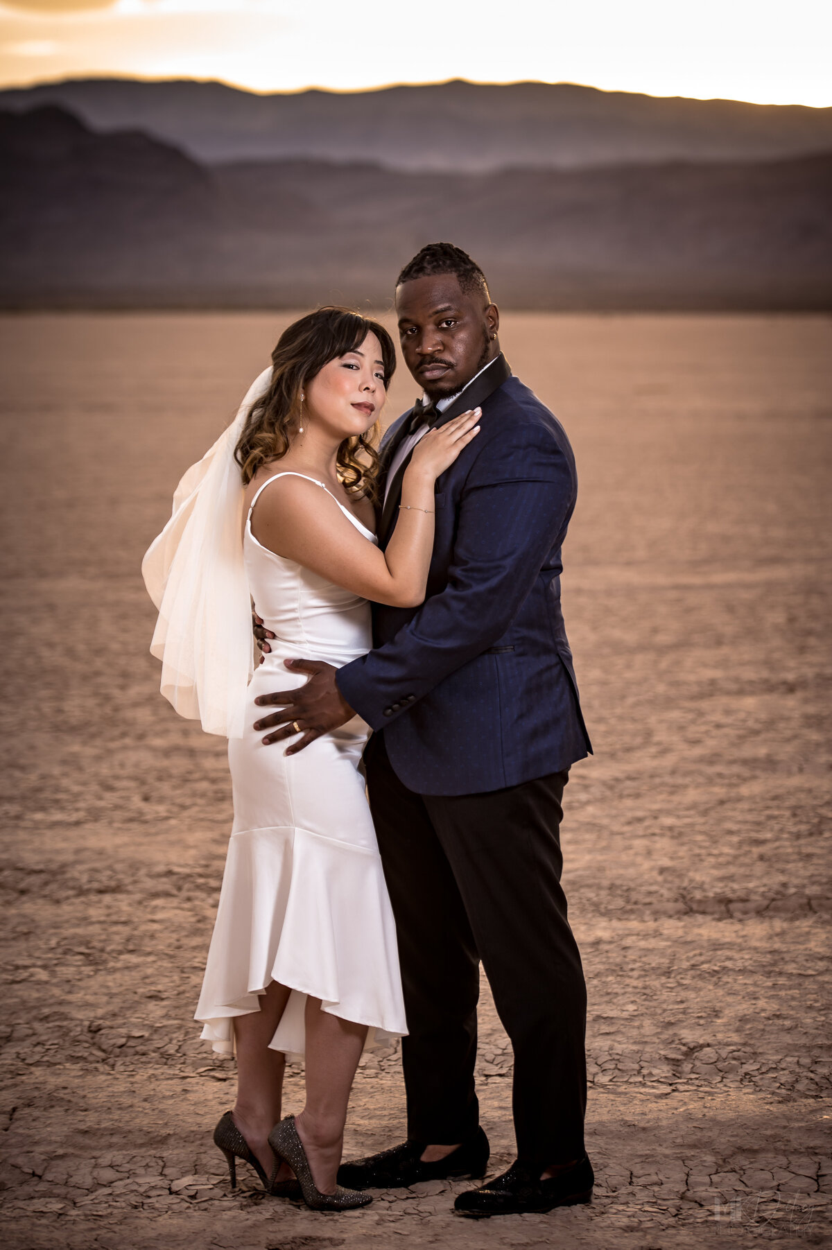 Military  veteran and bride bride and groom look at camer for their first portrait as husband and wife las vegas elopement on the dry lake bed  at golden hour groom in blue suit jacket and black  pants  las vegas elopement eloping in vegas  las vegas wedding photographers las vegas wedding photography mk delacy photography